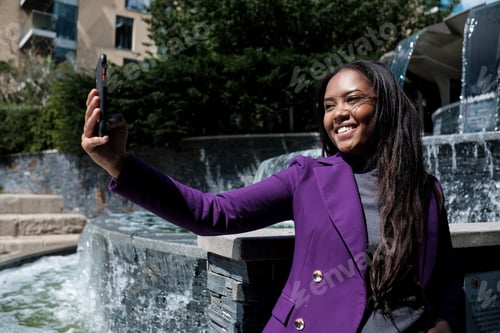 Preview: A beautiful young mixed race woman is taking a selfie in front of a fountain in a summer day.