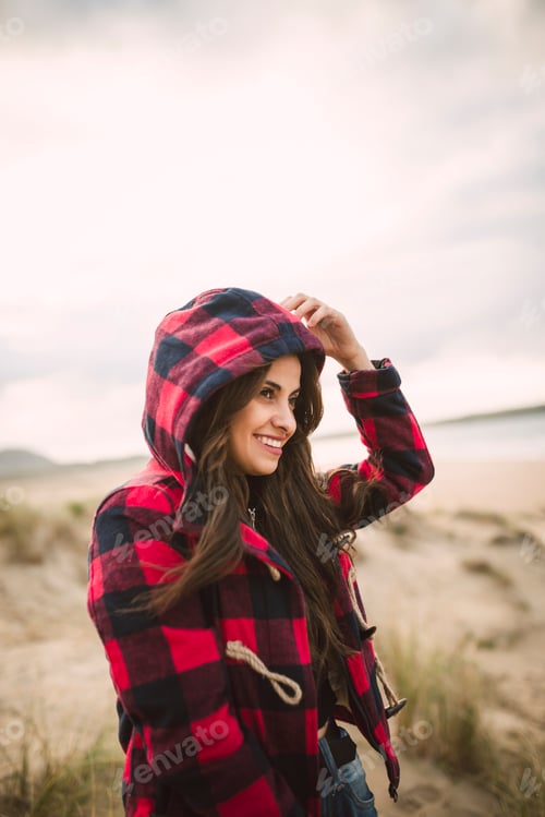 Preview: Smiling young woman with long brown hair wearing hooded jacket on the beach