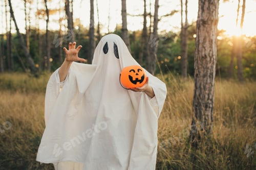Preview: Girl wearing ghost costume holding pumpkin bucket with candies, standing in a forest.