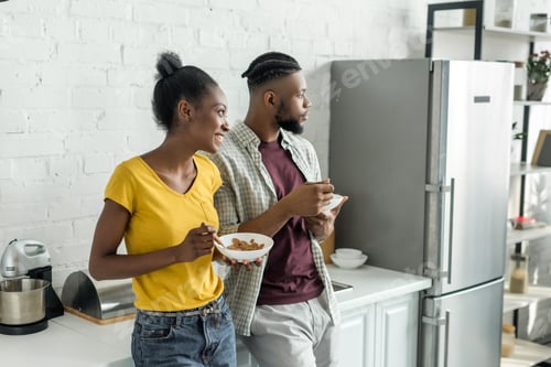 Preview: smiling african american couple having breakfast at kitchen and looking away
