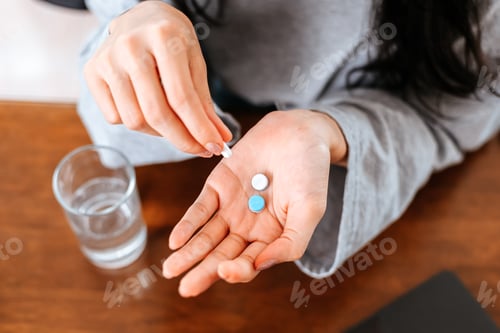 Preview: Businesswomen working at home with glass of water takes white round pill in hand.