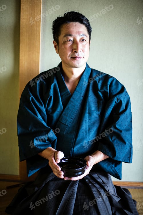 Preview: Japanese man wearing kimono kneeling on floor, holding tea bowl during tea ceremony.