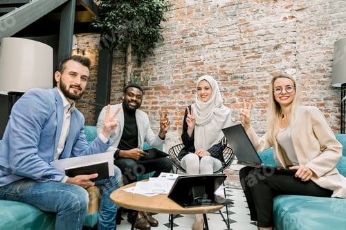 Preview: Happy cheerful multi-ethnic business team, two women and two men, showing victory hand sign looking