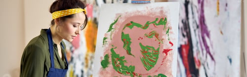 Preview: Creative young woman in apron painting abstract picture, applying paint on canvas with fingers