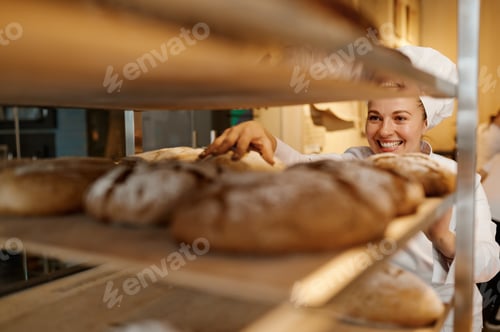 Preview: Happy woman baker in uniform checking freshly baked bread