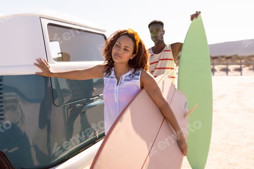 Preview: Front view of young Mixed-race couple with surfboard leaning on camper van at beach