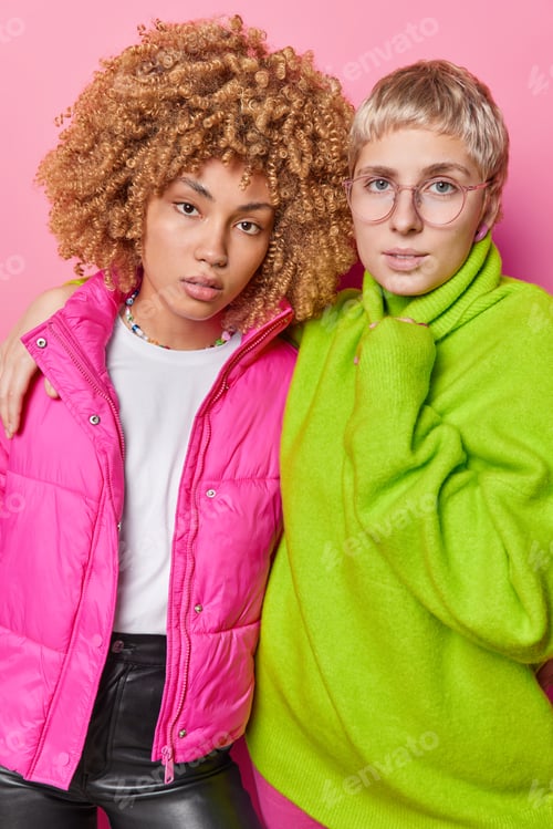 Preview: Two serious women embrace and stand closely to each other dressed in bright stylish clothed look