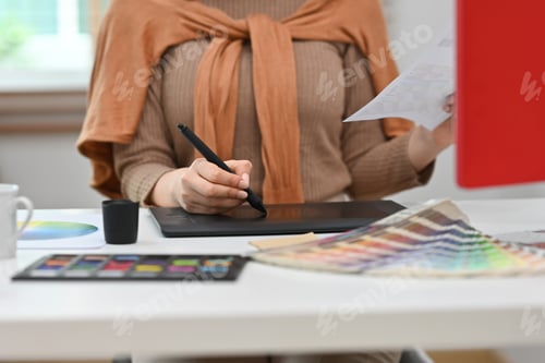 Preview: Young creative woman using graphic tablet and working with color swatch samples at workstation.