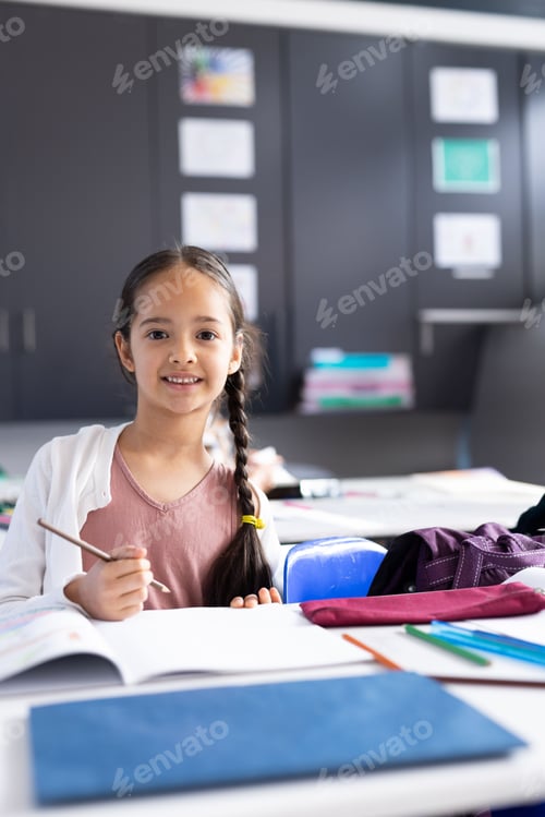 Preview: Female child sitting at desk in school classroom, holding pencil with workbook and colored pencils