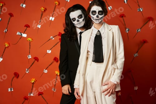 Preview: couple in dia de los muertos catrina makeup and suits looking at camera on red backdrop with flowers