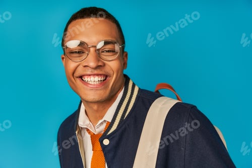 Preview: pleased african american student in eyeglasses and trendy jacket smiling at camera on blue, portrait