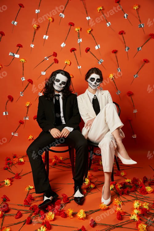 Preview: stylish couple in skull makeup and suits posing on chairs on red backdrop with flowers, Day of Dead