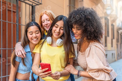 Preview: Four multiracial female friends using smartphone watching funny video on social media on the street
