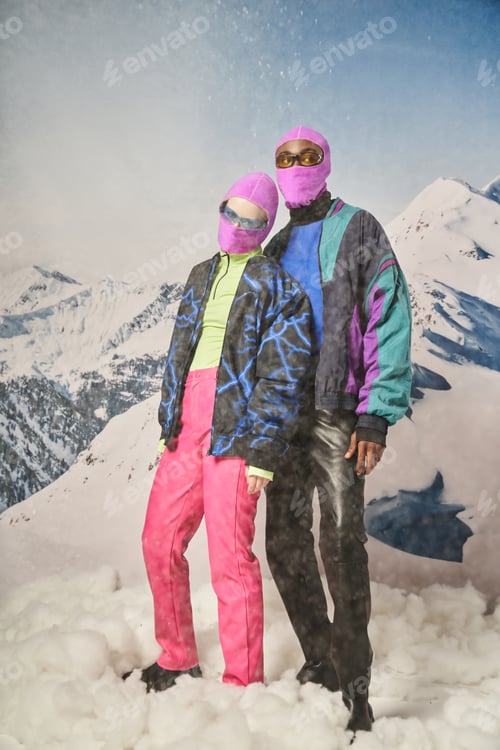 Preview: attractive diverse couple posing together in pink balaclavas with mountain backdrop, winter concept