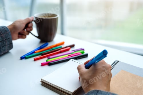 Preview: Closeup image of a woman writing on a blank notebook with colored pens while drinking coffee