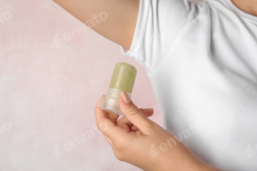 Preview: Young woman holding natural crystal alum deodorant near armpit on light pink background, closeup