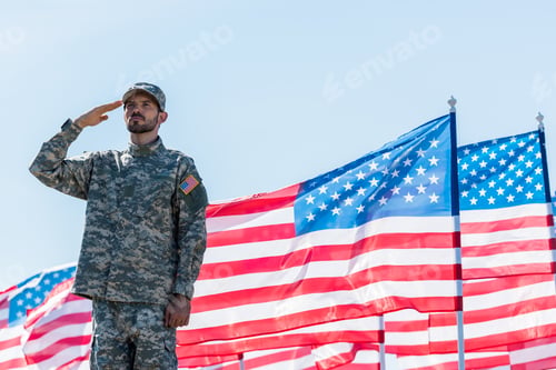 Preview: handsome soldier in military uniform giving salute near american flags with stars and stripes