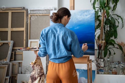 Preview: Rearview focused female artist completes last brushstrokes on sky sea landscape painting in studio