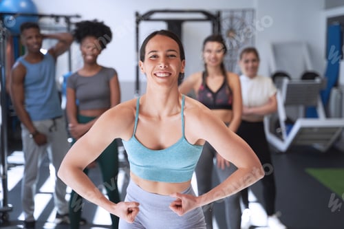 Preview: Woman showing her arm muscle while posing in front of a group at the gym.