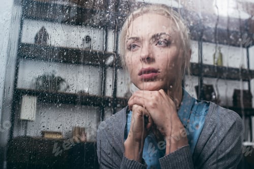Preview: sad adult woman with folded hands looking away at home through window with raindrops