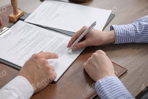 Preview: Notary pointing at document and client putting signature at wooden table indoors, closeup