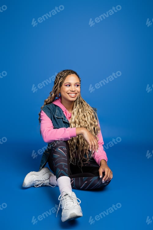 Preview: full length of young long haired african american woman sitting in sporty outfit on blue background