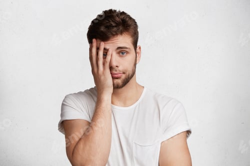 Preview: What pity! Attractive blue eyed unshaven male with trendy hairdo, covers face with hand as expresses