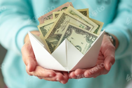 Preview: Hands hold a paper boat filled with banknotes in a simple indoor setting