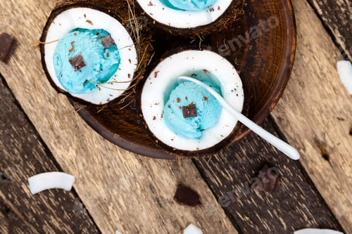 Preview: Blue ice cream with chocolate in coconut bowl on wooden background.