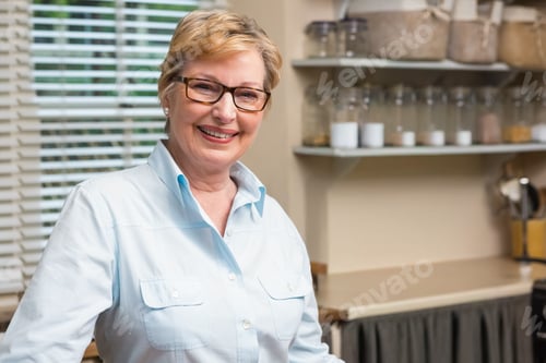 Preview: Senior woman standing and smiling in kitchen at counter, wearing blue shirt and glasses