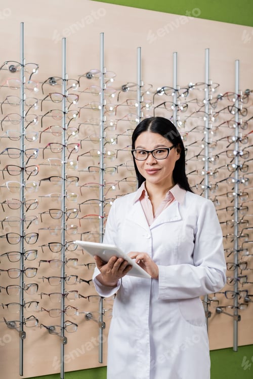 Preview: happy asian oculist looking at camera while using digital tablet in optics salon