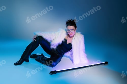 Preview: trendy brunette woman in white faux fur jacket looking at camera near fluorescent lamp on blue