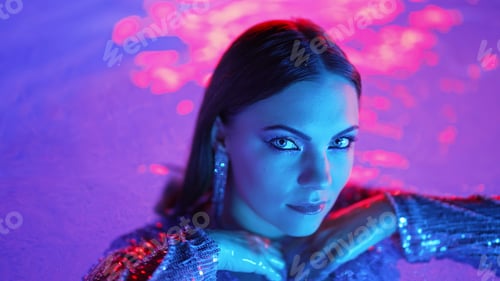 Preview: Beautiful woman posing in pool water under neon color light. Party, attractive chic in shining dress