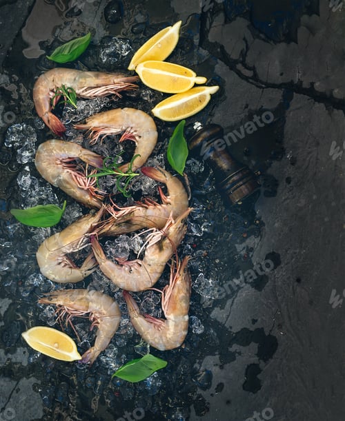 Preview: Fresh uncooked shrimps with lemon, herbs and spices on chipped ice over dark slate stone backdrop