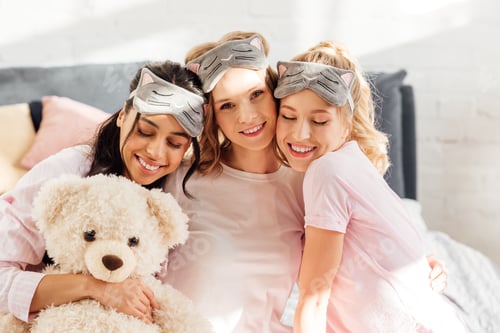 Preview: beautiful happy multicultural girls in sleeping masks hugging in morning during pajama party