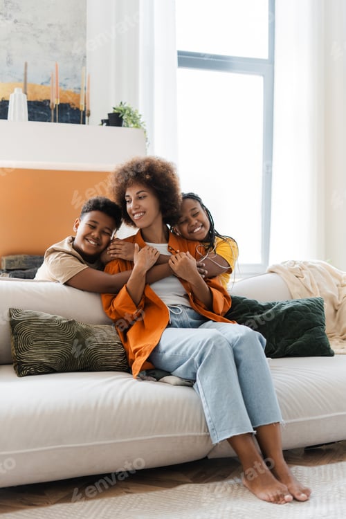Preview: Funny African American children enjoying, embracing with their mother, sitting on sofa