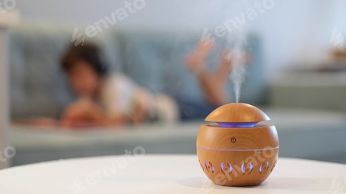 Preview: Modern aroma oil diffuser on the white table. Little boy in the background reading a book