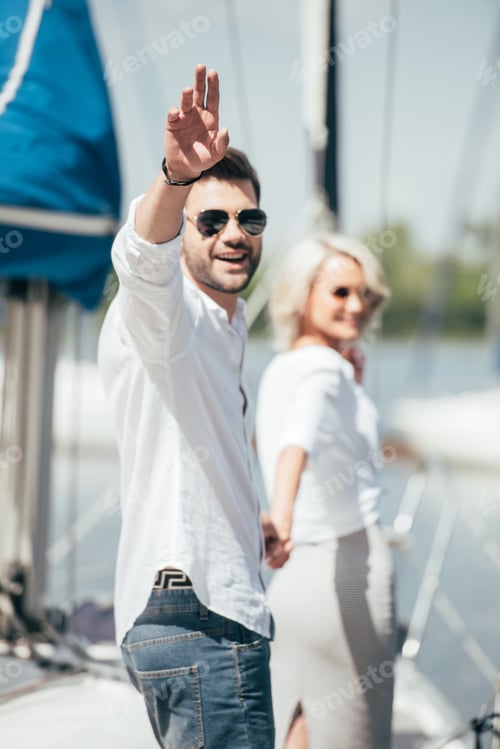 Preview: young man in sunglasses smiling at camera and waving hand while holding hands with beautiful girl on