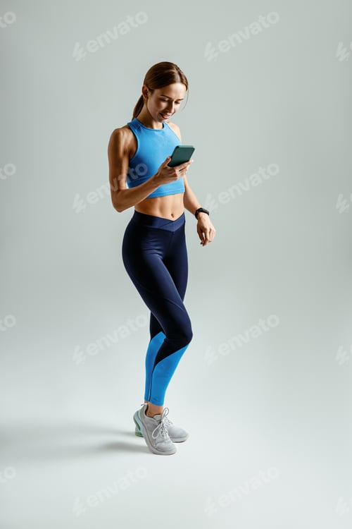 Preview: Athletic woman in costume for workout holding smartphone standing against studio background