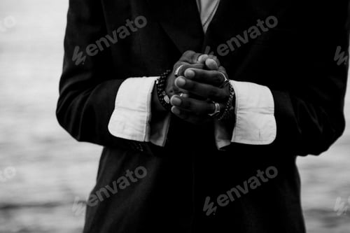 Preview: Grayscale shot of a black man wearing a coat with a white shirt, hands clasped in the front