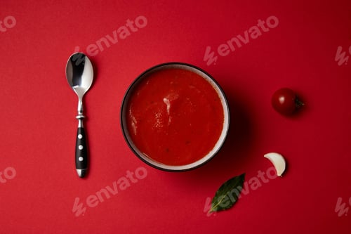 Preview: top view of tasty tomato soup in plate, spoon, fresh tomato and garlic on red table