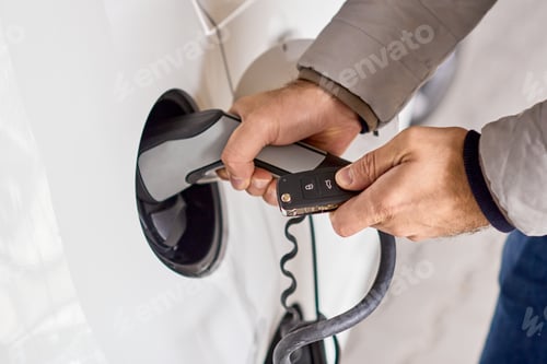Preview: Closeup mans hand holding electric car charging connected to electric car at public charging station