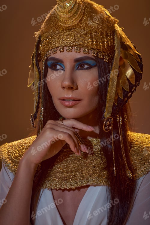 Preview: Portrait of stylish woman in egyptian headdress and bold makeup posing isolated on brown