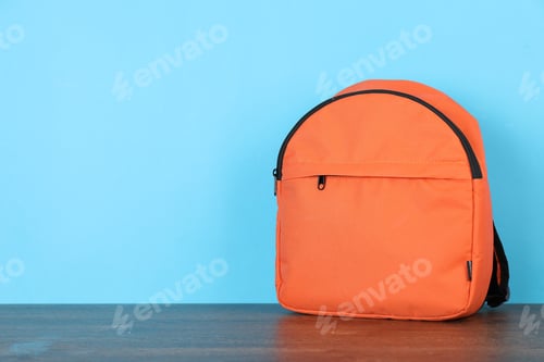 Preview: Orange backpack on wooden table against light blue background. Space for text