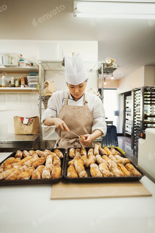 Preview: Woman wearing chef's hat and apron working in a bakery, preparing freshly baked rolls on large