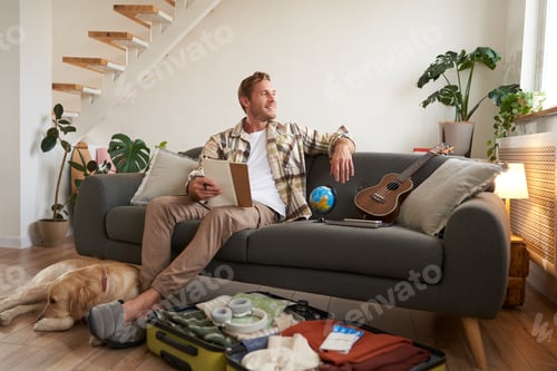 Preview: Portrait of young man sitting on sofa with notebook, has dog lying next to him, packing suitcase