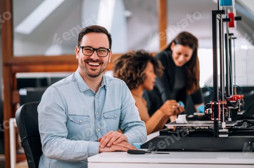 Preview: Portrait of a smiling entrepreneur at work. Working with 3D printer, coworkers in the background.