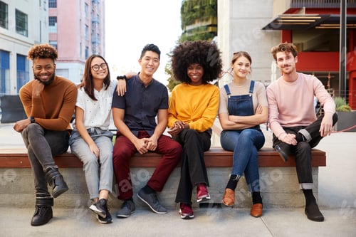 Preview: Six young adult friends sitting in a row on a bench in a city street smiling to camera, full length