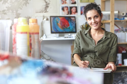 Preview: Positive young creative woman dressed casually, sitting at her workshop, making sketches with pencil
