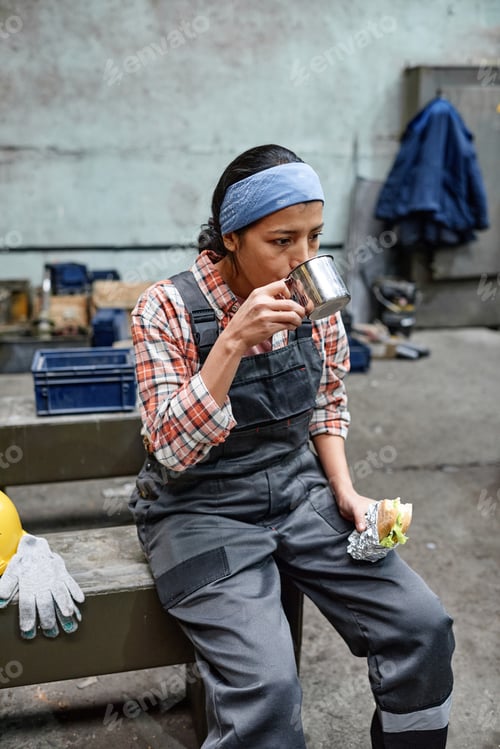 Preview: Young tired and hungry female engineer having tea with sandwich at break
